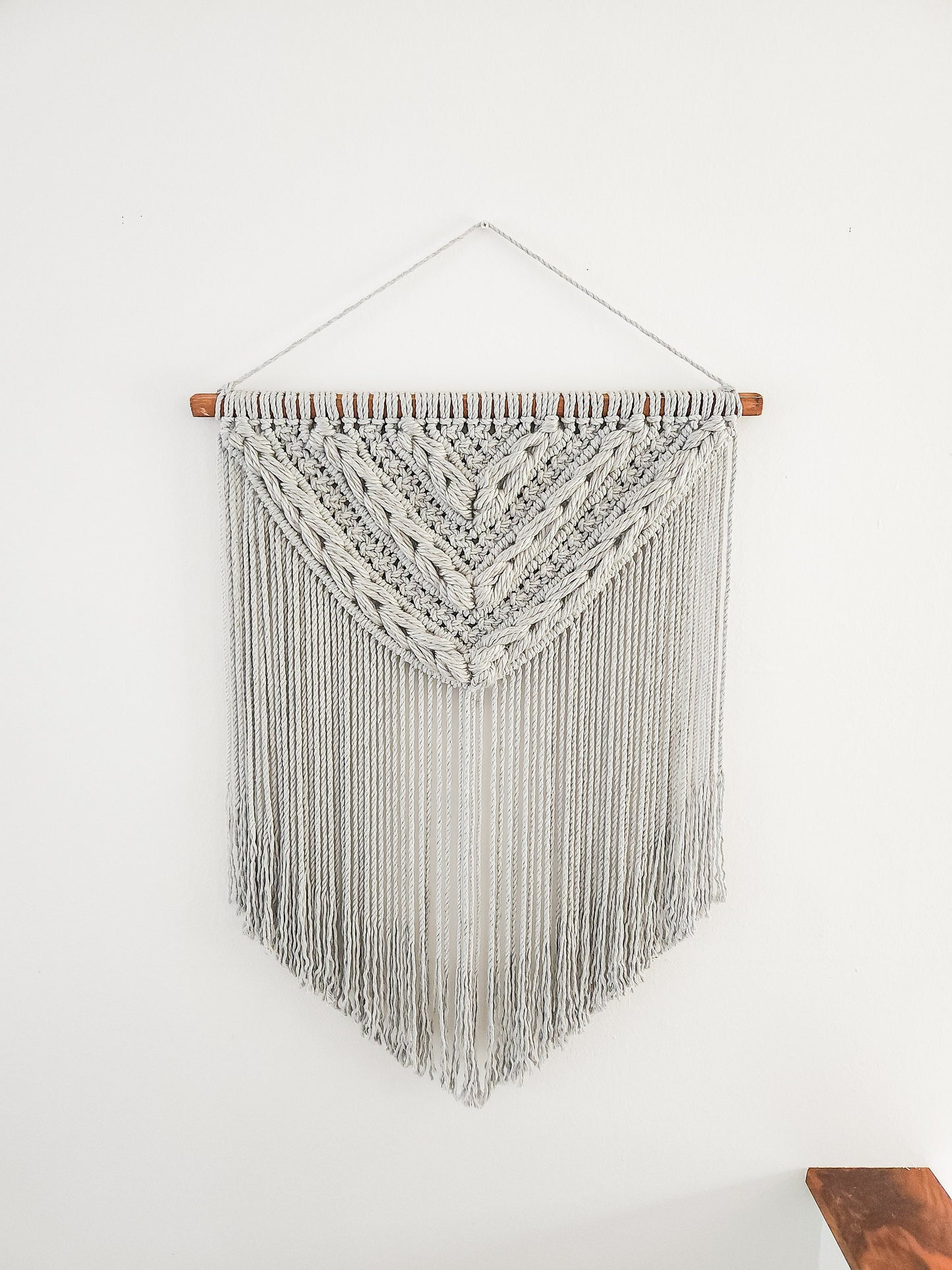 Light gray macrame wall hanging with light gray woven section. Shown on a white wall background.