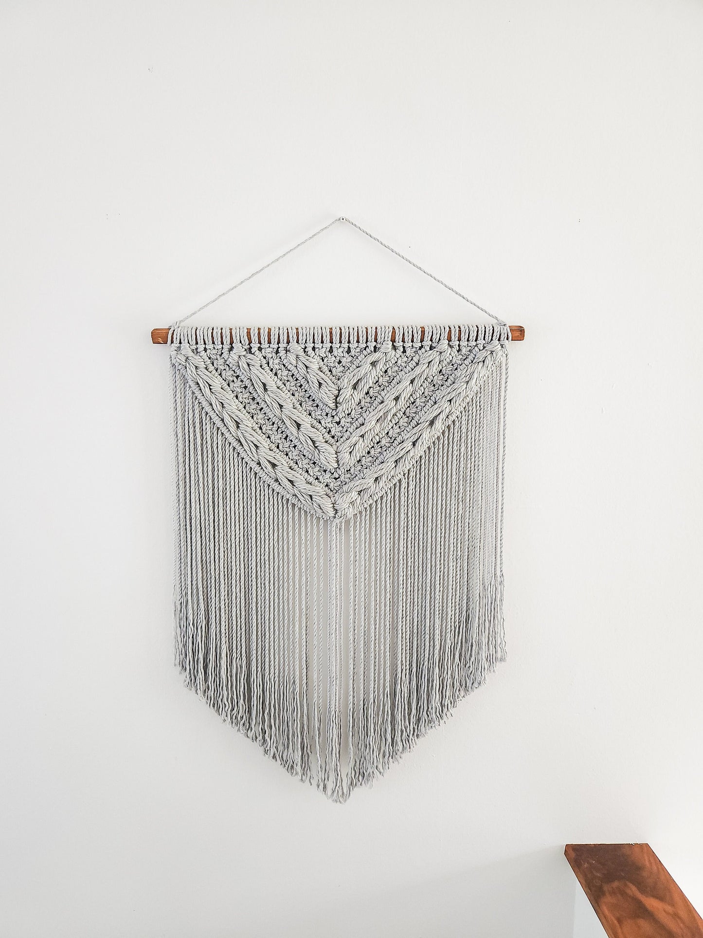 Light gray macrame wall hanging with light gray woven section. Shown on a white wall background.