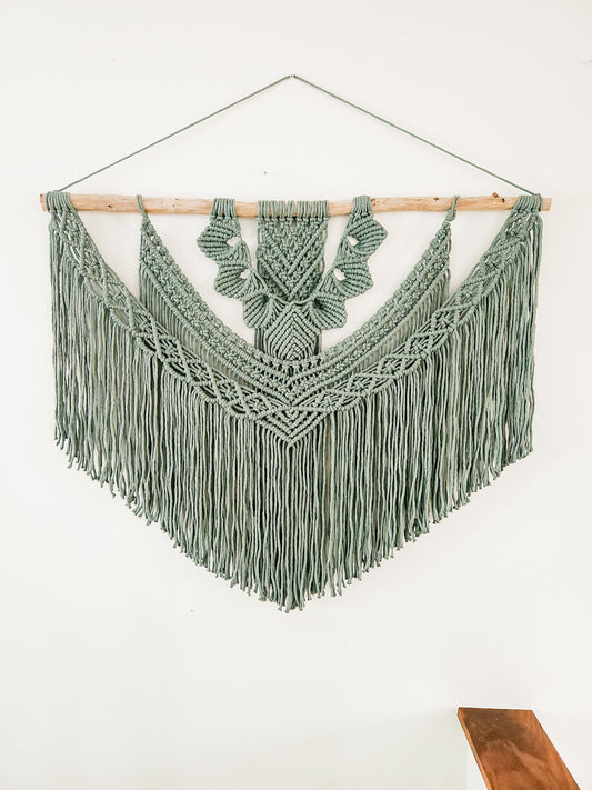 Extra large macrame wall hanging on a piece of driftwood. The wall hanging is in a laurel color and it is shown on a white wall.
