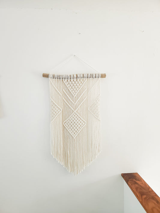 Beige macrame wall hanging on driftwood on a white wall.