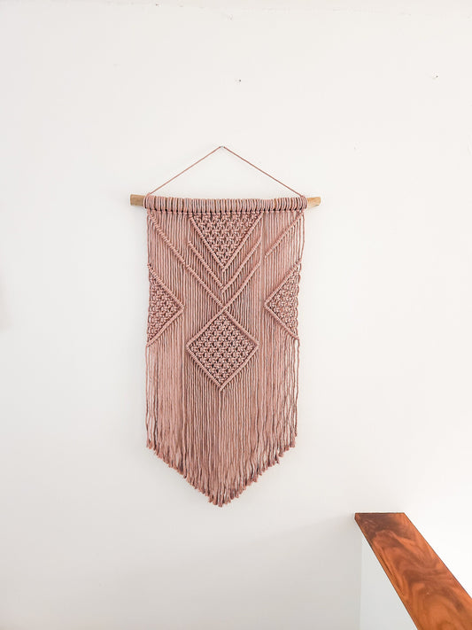 Mauve colored macrame wall hanging on driftwood. Shown on a white wall.