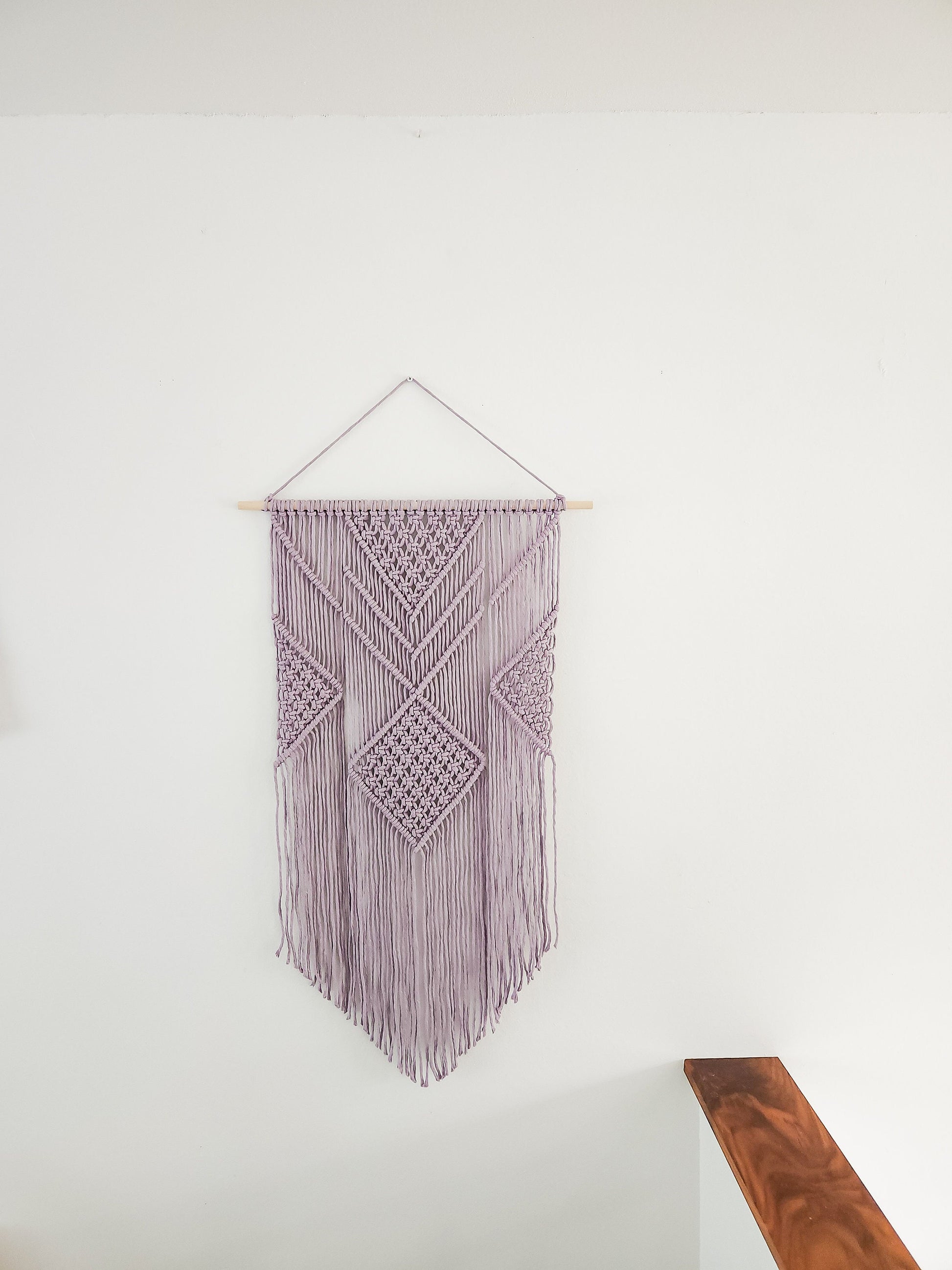 Lavender purple macrame wall hanging on a white wall. It is 21.5 inches wide and 36 inches long, Aztec style design.