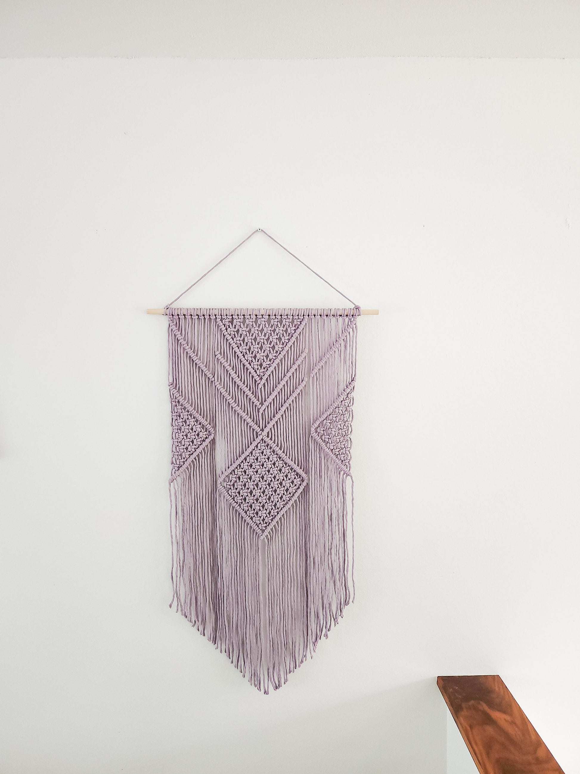 Lavender purple macrame wall hanging on a white wall. It is 21.5 inches wide and 36 inches long, Aztec style design.