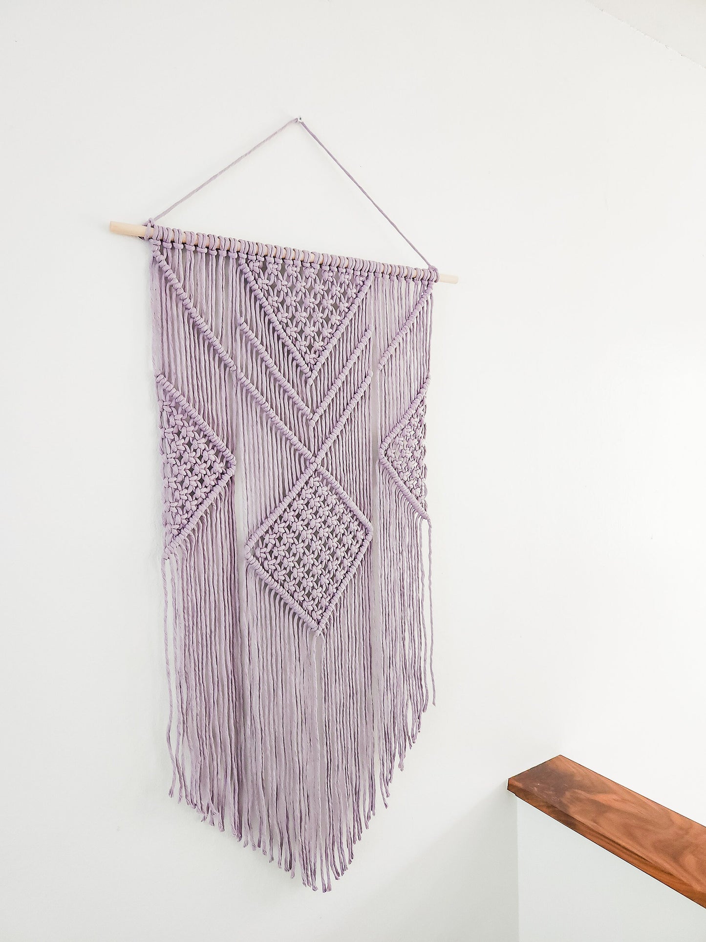 Lavender purple macrame wall hanging on a white wall. It is 21.5 inches wide and 36 inches long, Aztec style design.