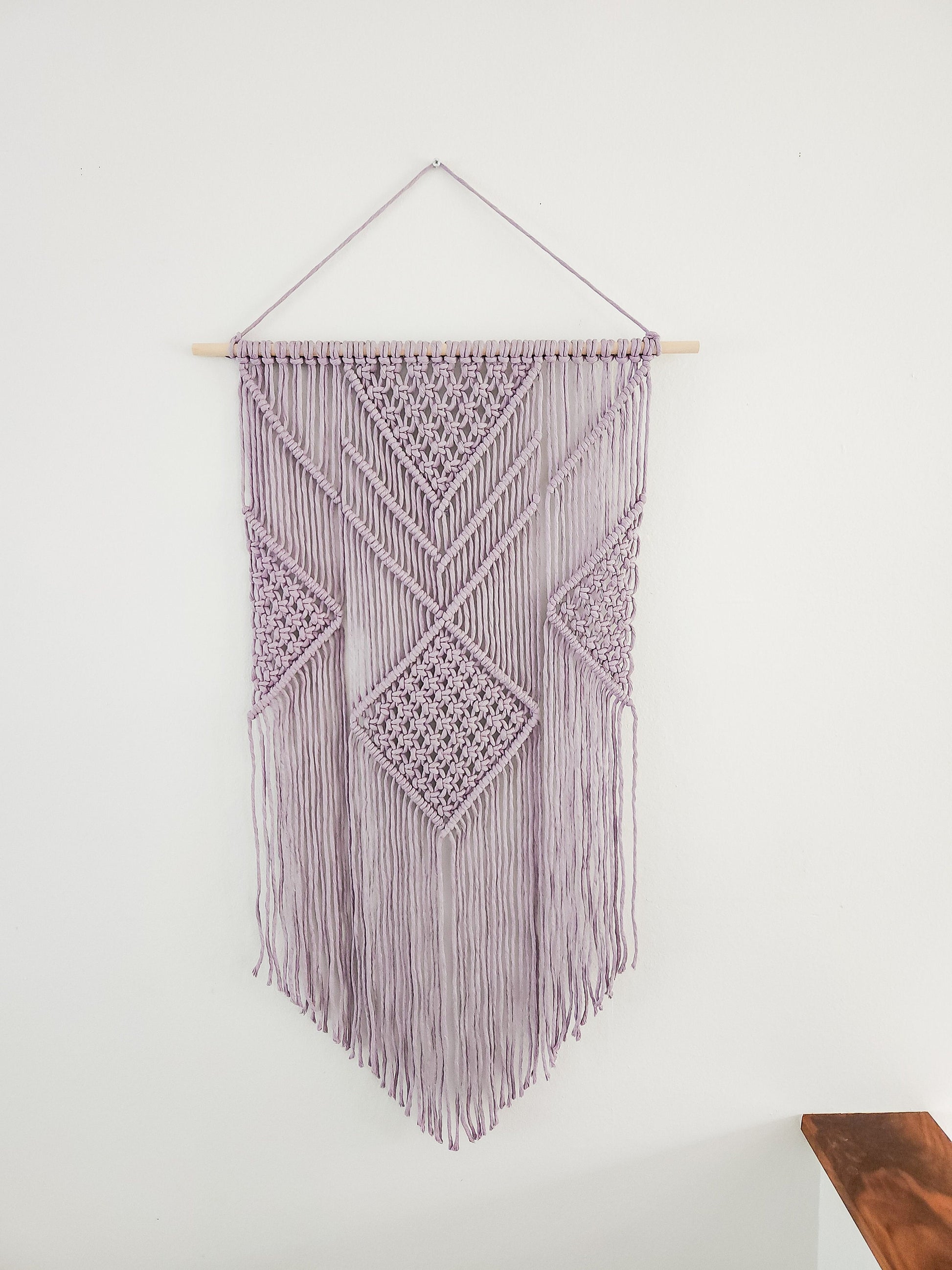 Lavender purple macrame wall hanging on a white wall. It is 21.5 inches wide and 36 inches long, Aztec style design.
