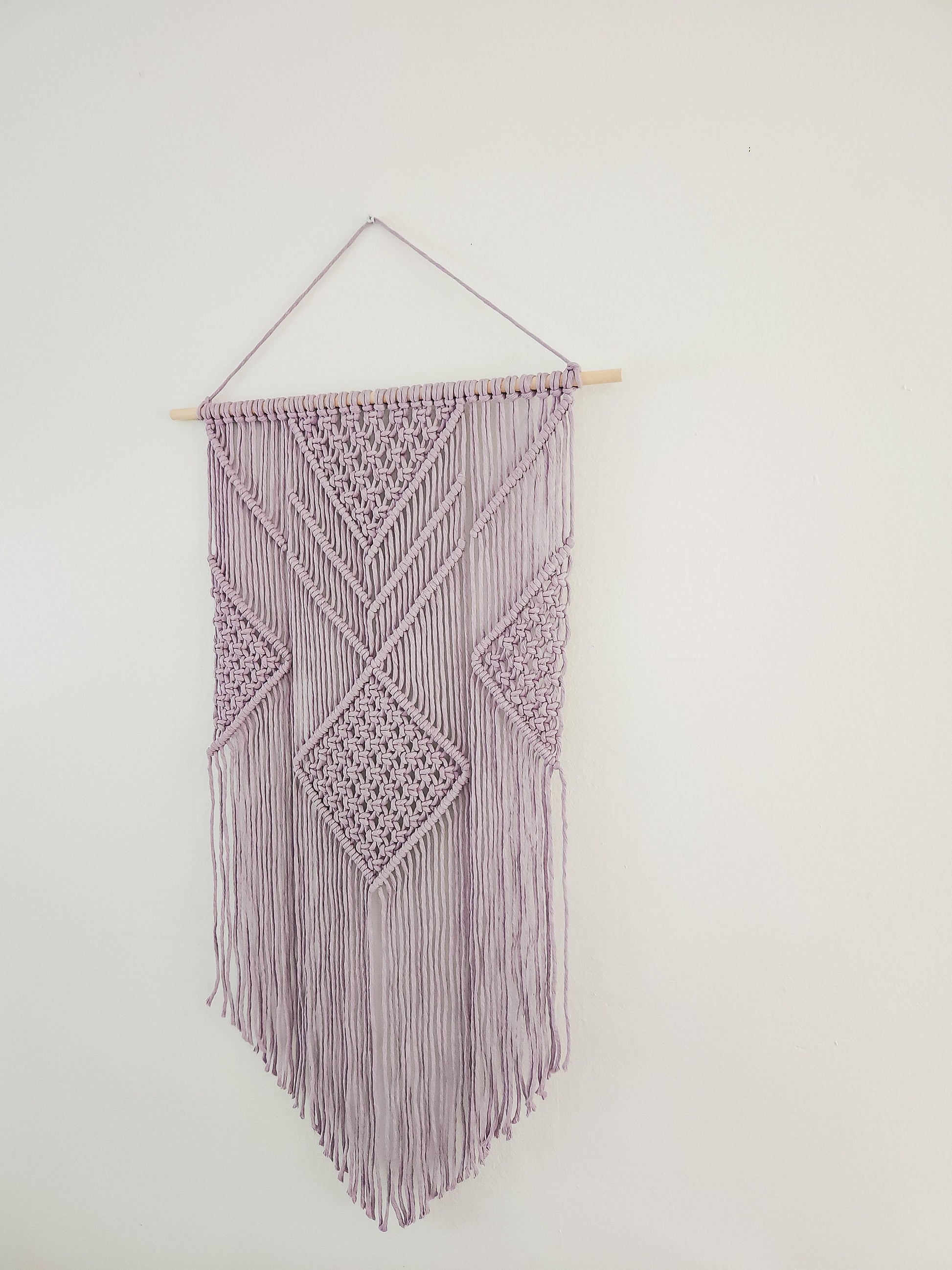 Lavender purple macrame wall hanging on a white wall. It is 21.5 inches wide and 36 inches long, Aztec style design.
