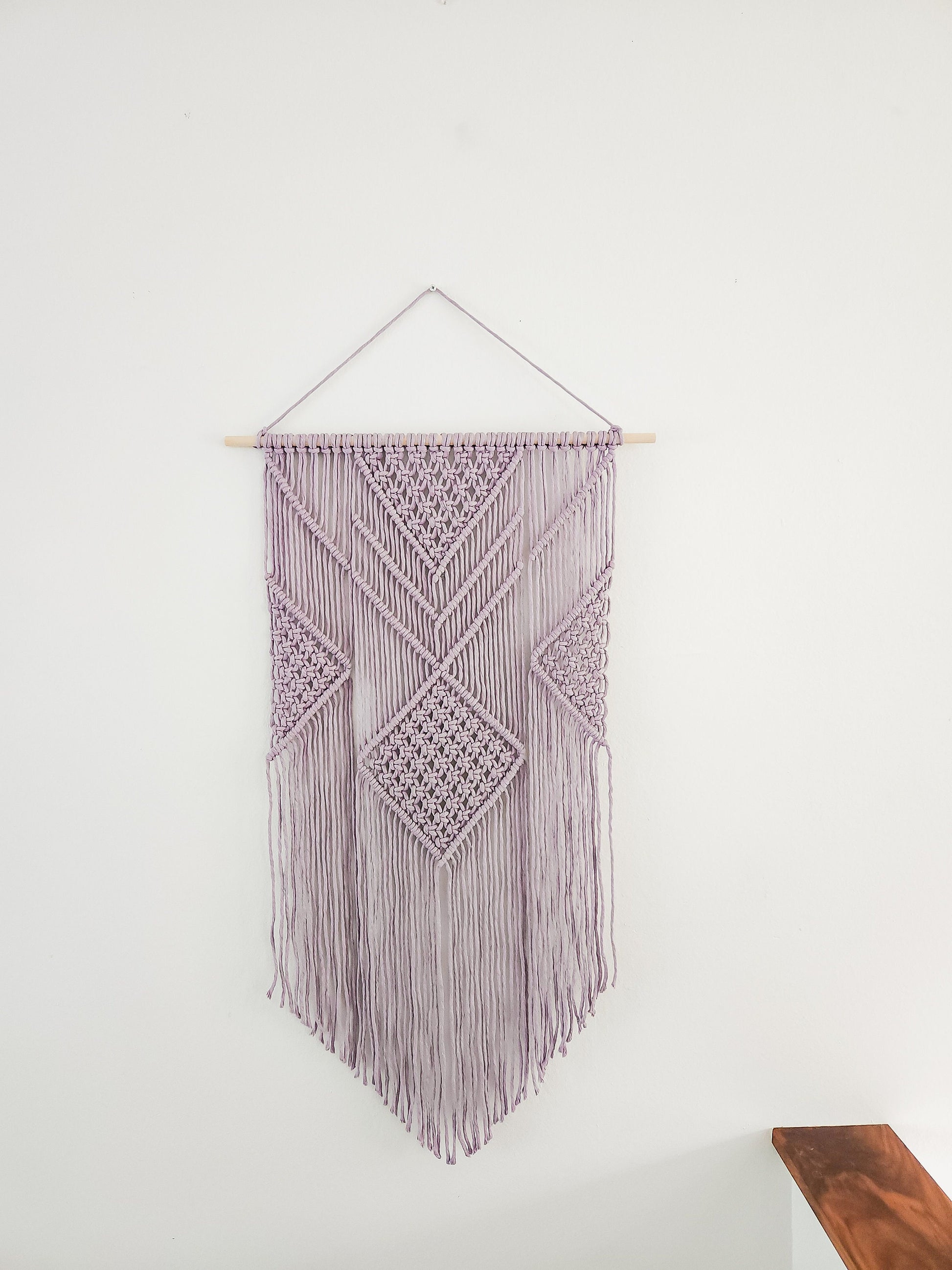 Lavender purple macrame wall hanging on a white wall. It is 21.5 inches wide and 36 inches long, Aztec style design.