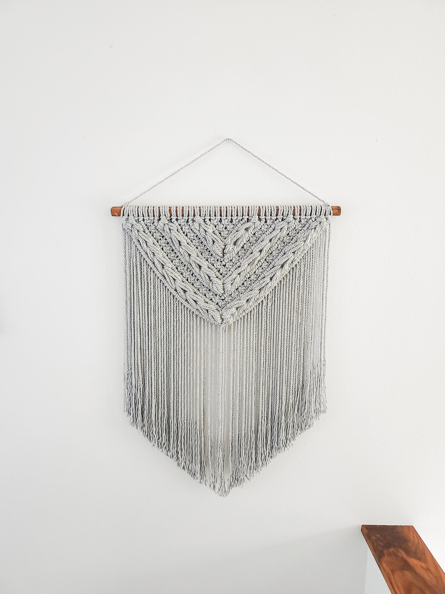 Light gray macrame wall hanging with light gray woven section. Shown on a white wall background.