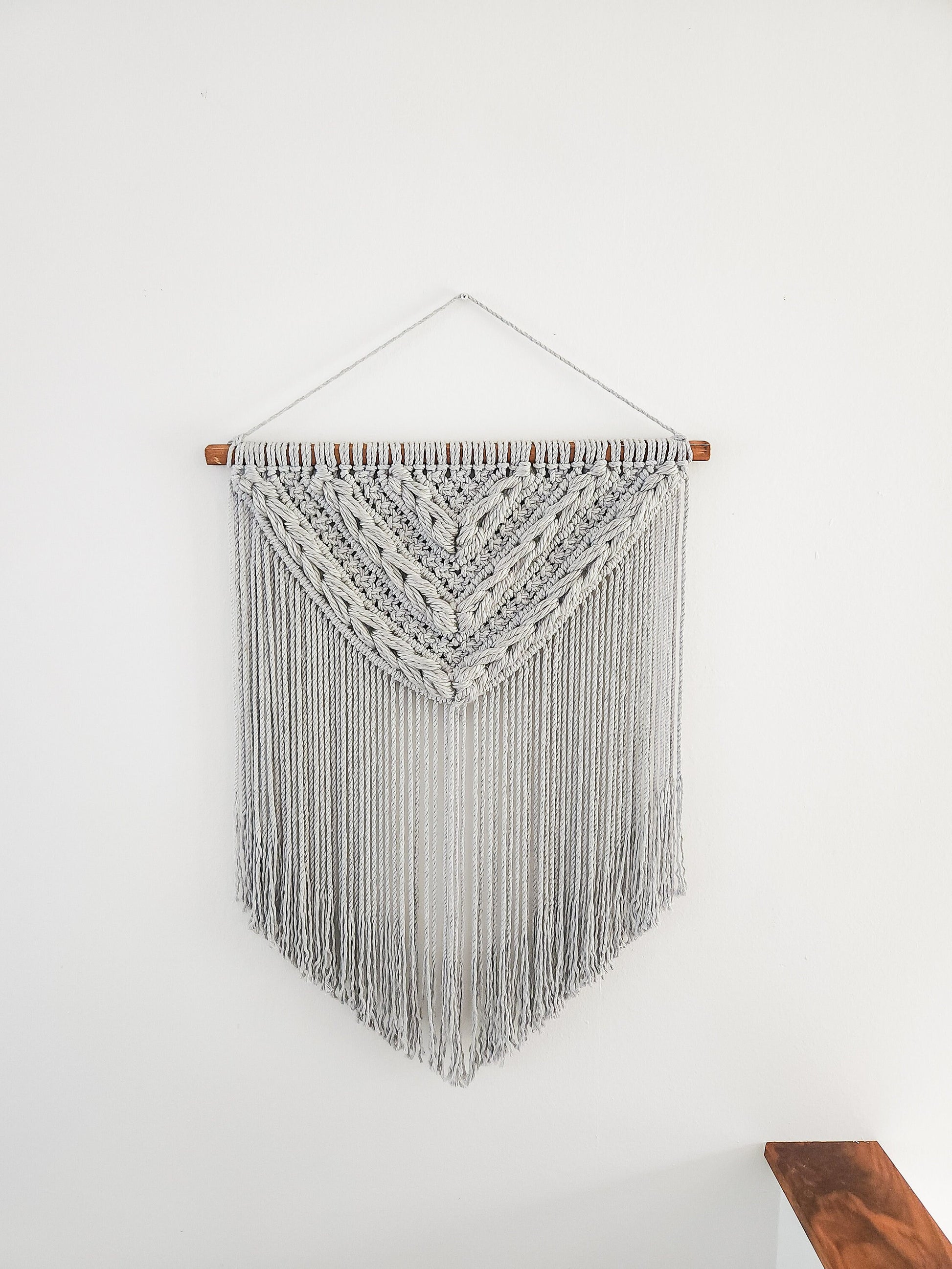 Light gray macrame wall hanging with light gray woven section. Shown on a white wall background.