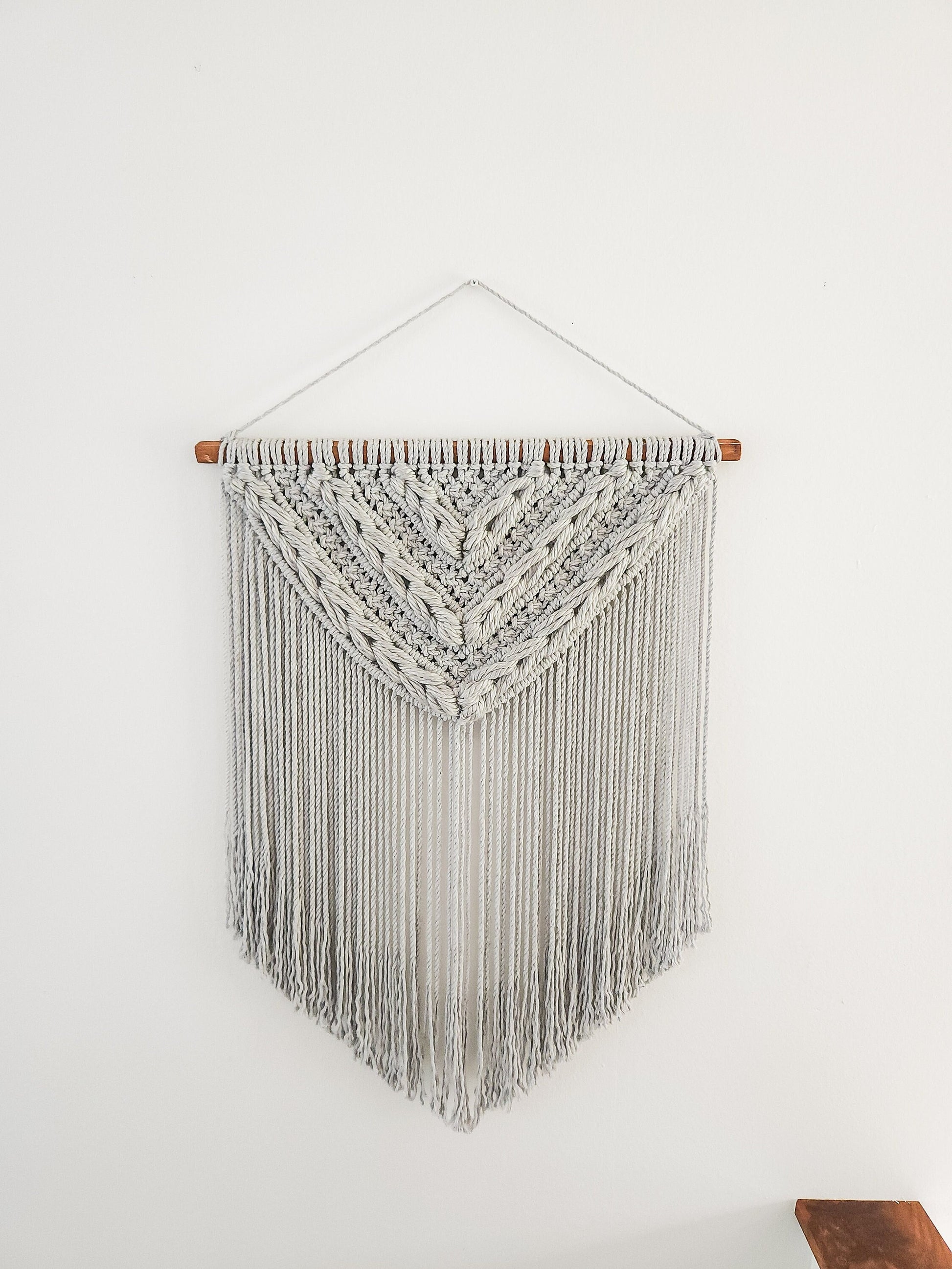 Light gray macrame wall hanging with light gray woven section. Shown on a white wall background.