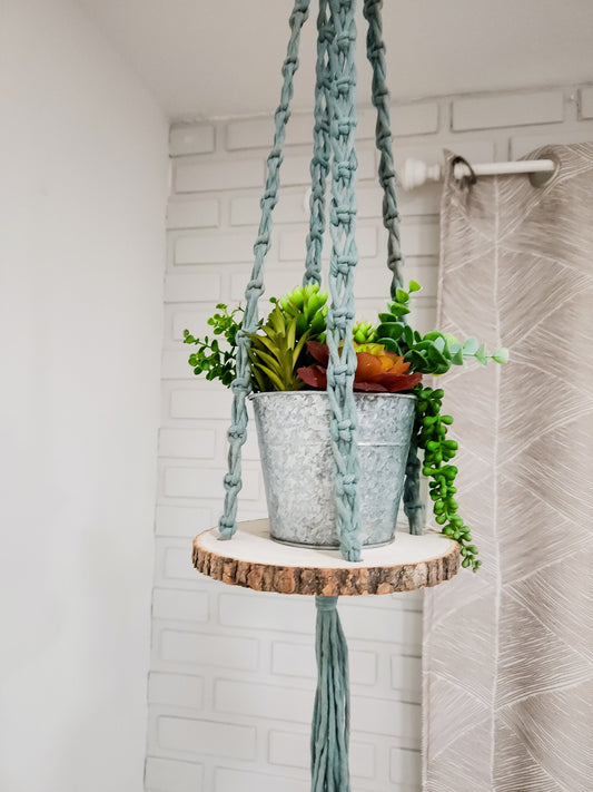 Macrame hanging shelf. It comes in a variety of cord colors, and it has a wood round with bark on the sides.